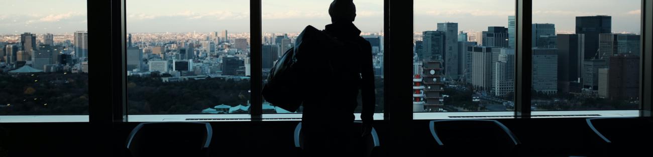 Homme de dos en contre-jour, un sac de sport à l'épaule, observant la ville à travers de grandes vitres