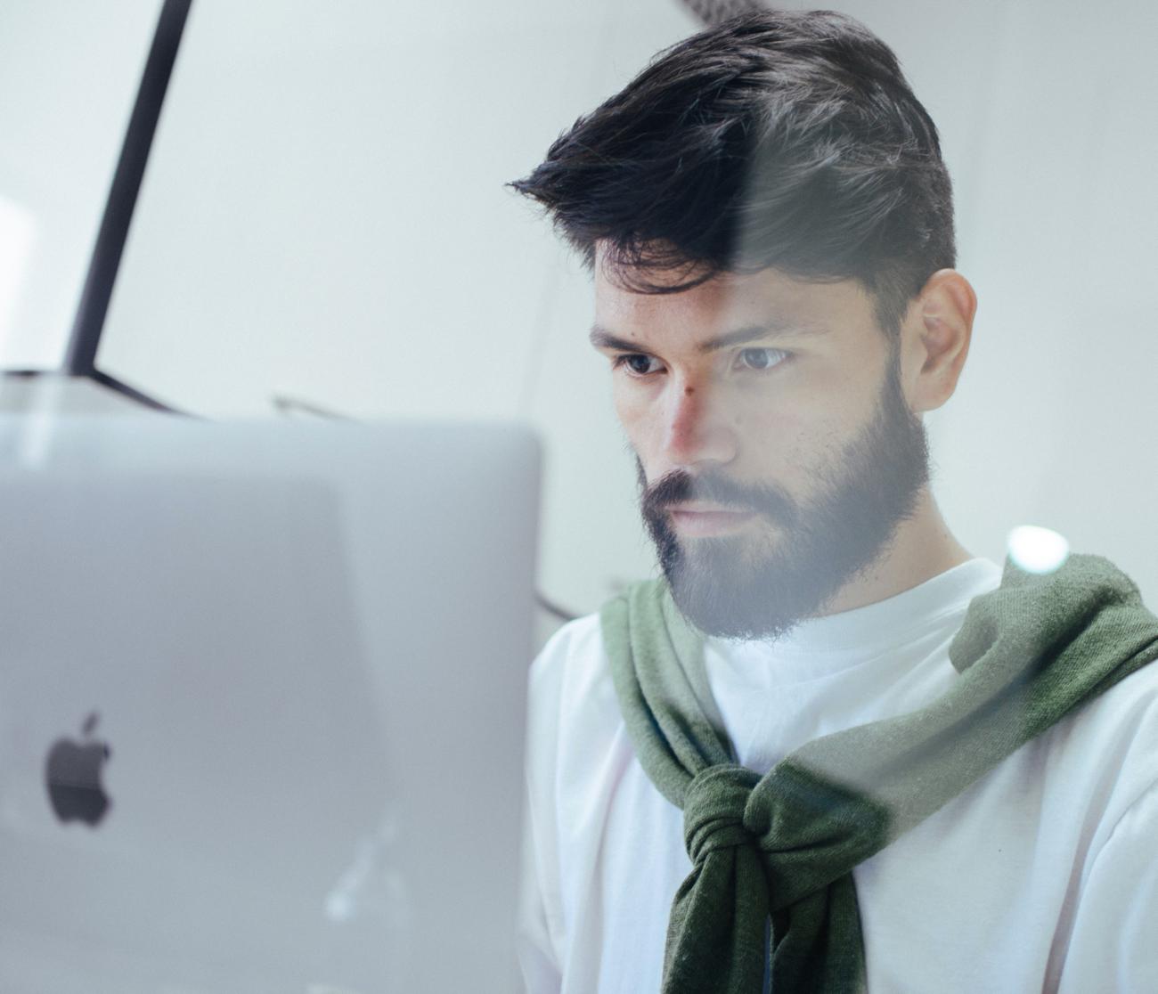 Homme avec brushing, barbe et t-shirt en bandoulière au niveau des épaules, regardant son écran d'ordinateur Mac
